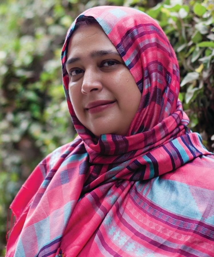 Author Saadia Faruqi looking at the camera with a slight smile, posed in front of a large green bush.